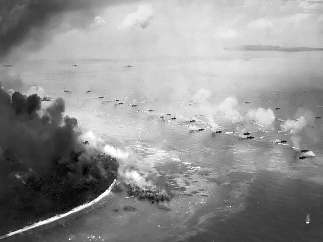 The first wave of LVTs moves toward the invasion beaches, passing through the inshore bombardment line of LCI gunboats. Cruisers and battleships are bombarding from the distance. The landing area is almost totally hidden
