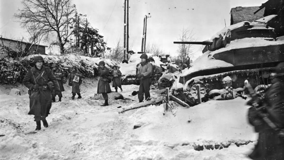 Members of the 117th Infantry Regiment, 30th Infantry Division, move past a destroyed American M5 "Stuart" tank on their march to capture the town of St. Vith at the close of the Battle of the Bulge. St. Vith was largely