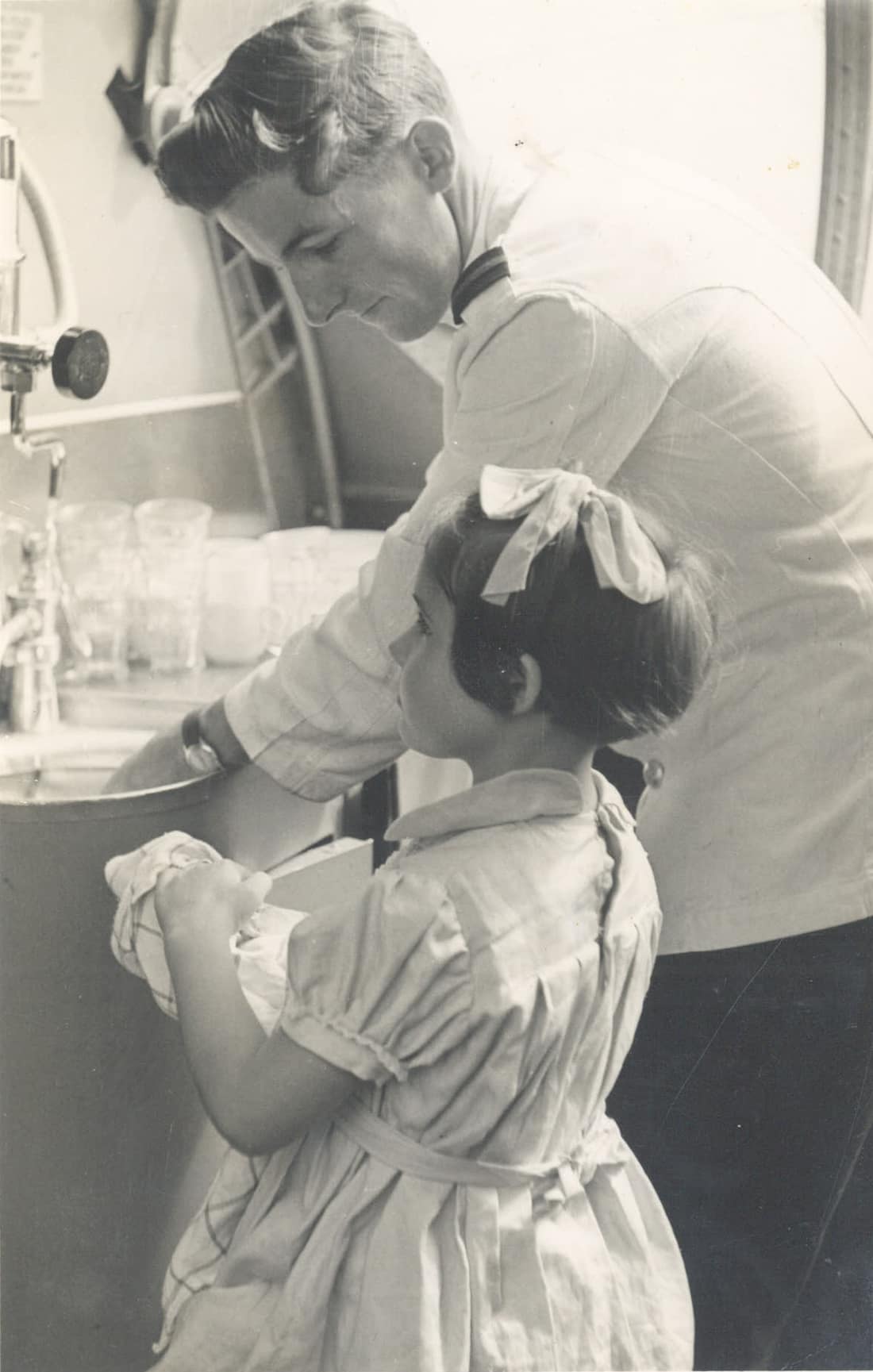 Flight steward Max White at work on board a Qantas Catalina aircraft en route from Suva to Sydney in January 1949, assisted by young passenger Jennifer Grey.