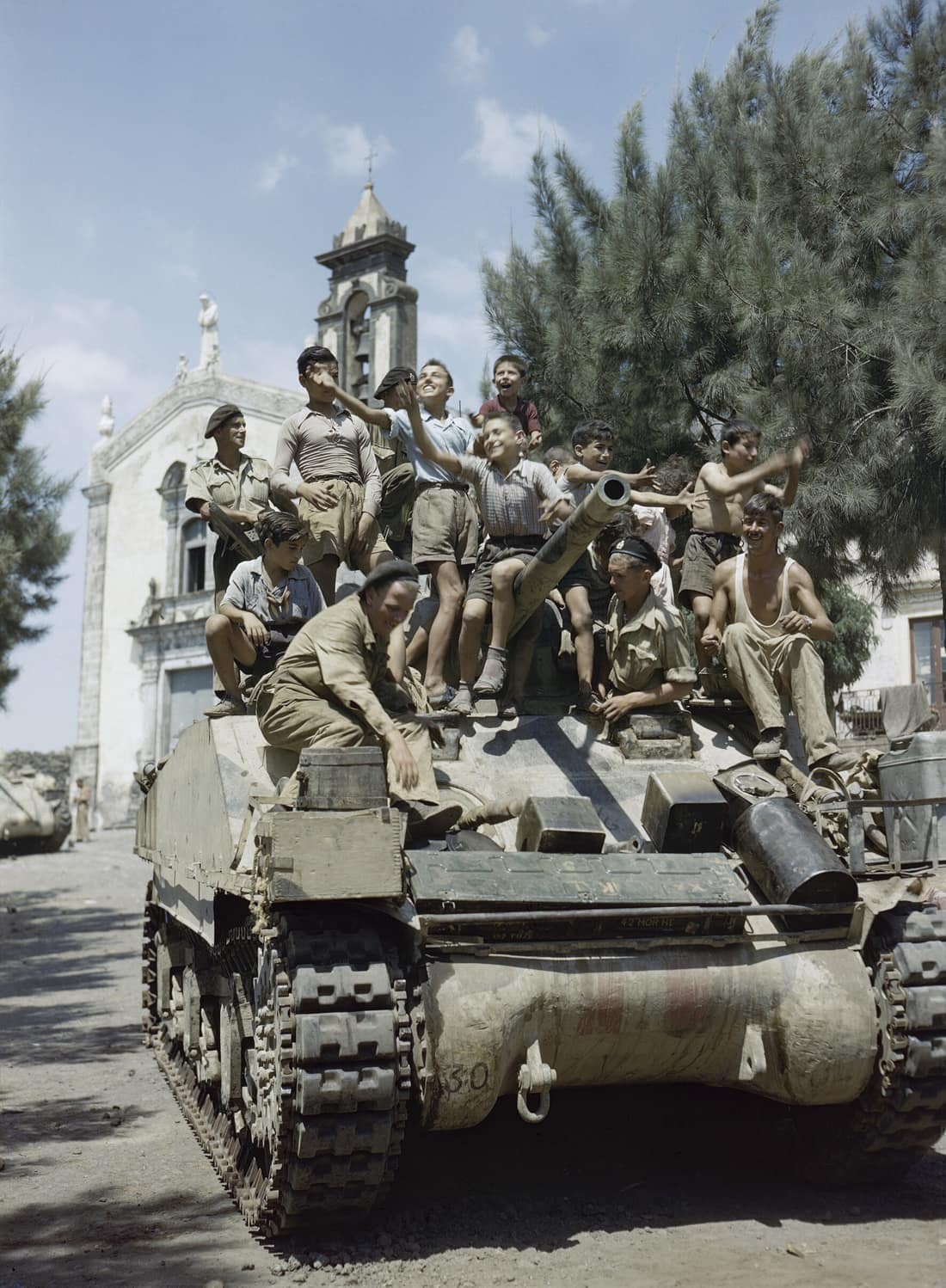 Local children hitch a ride on a Sherman tank of 3rd County of London Yeomanry (Sharpshooters) in Belpasso near Catania, Sicily, August 1943. In the background can be seen the church of Saint Anthony of Padua. At Belpass
