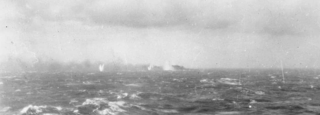 The German battleship Bismarck on fire in the distance, surrounded by shell splashes. The photo was taken from one of the Royal Navy warships chasing.